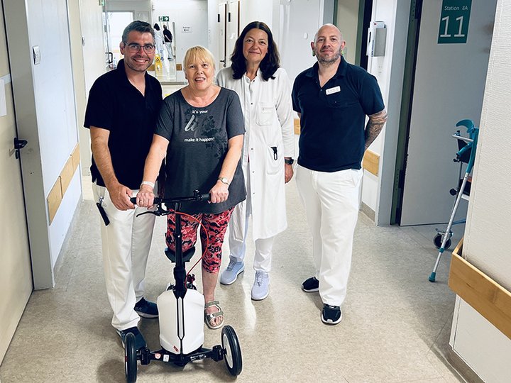 Frau auf einem Spezialroller im Krankenhaus, begleitet von Ärztin und zwei Physiotherapeuten