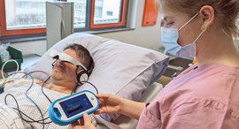 Person liegt im Bett und hat eine Videobrille an. Eine andere Person steuert das Gerät am Computer