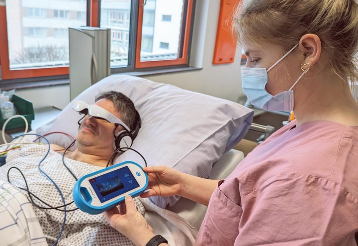 Person liegt im Bett und hat eine Videobrille an. Eine andere Person steuert das Gerät am Computer