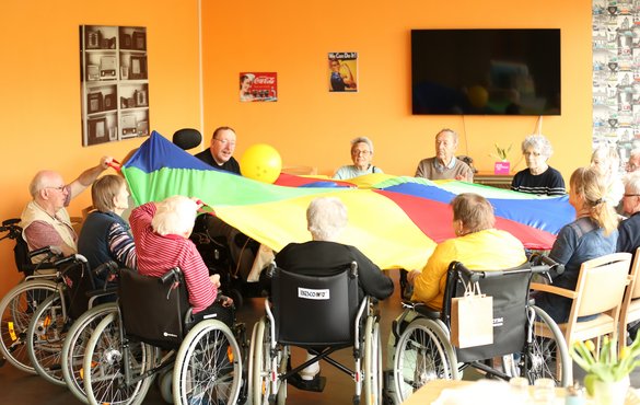 Seniorengruppe im Sitzkreis balanciert einen Ball auf einem großen Regenbogentuch