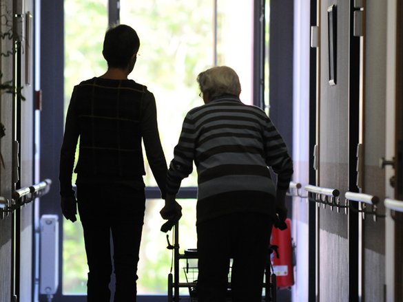 Rückenansicht einer Seniorin am Rollator in Begleitung in einem Flur 