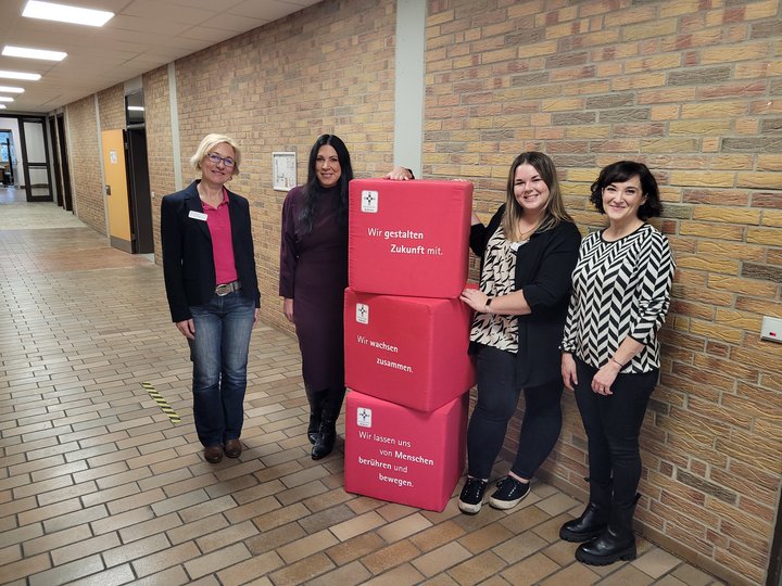 Vier Frauen stehen im Gang der Schule. Zwischen ihnen sind rote Würfel.