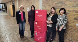 Vier Frauen stehen im Gang der Schule. Zwischen ihnen sind rote Würfel.